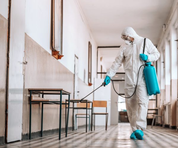 Worker in sterile white uniform, with mask and glasses holding sprayer with disinfectant and spraying around hallway in school. Prevention of spreading corona virus.